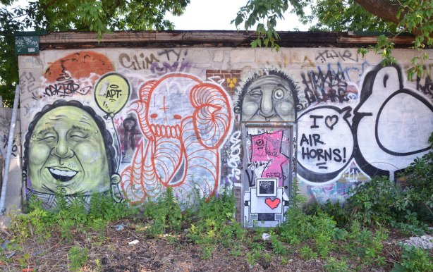 a wall covered with graffiti, a large bald man's head, a red and white elephant, a lovebot, a black line drawn bird saying I love air horns