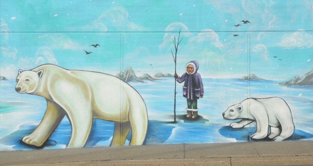arctic scenes as part of a larger street art mural on an underpass in Toronto - a person in a purple parka holding a little tree, standing in ice between two polar bears
