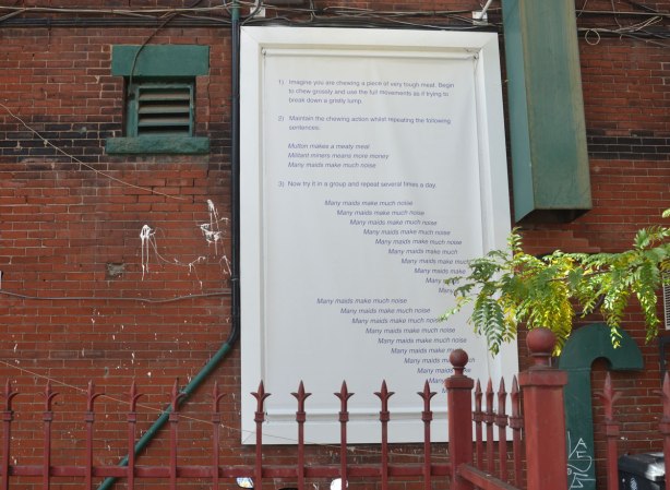 words in dark blue written on a white board that is fastened to a brick wall, behind a rust coloured metal fence