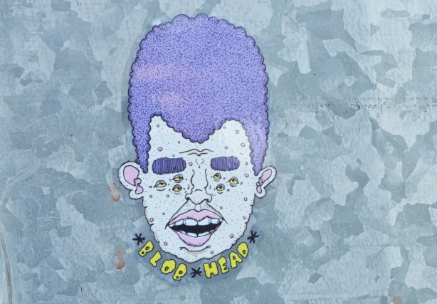 sticker on a grey metal pole. head with the words Blob Head written underneath. Purple hair, open mouth, pink lips, teeth showing, purple bushy eyebrows and three small eyes where each eye should be (six eyes in total)