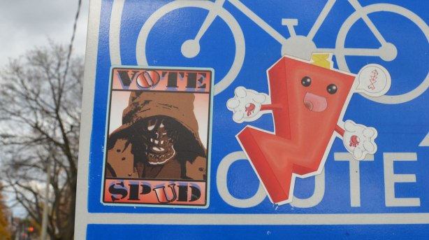 close up of two stickers on a blue and white bike route sign. One sticker is a brown one with the words Vote Spud and a picture of skull wearing a floppy hat. The other is a red cartoon character 