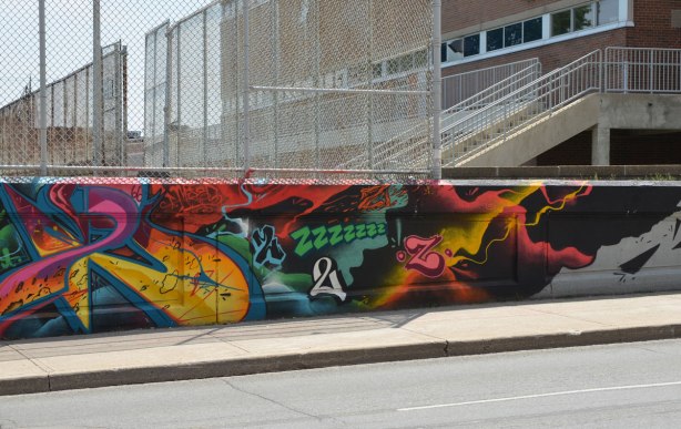 wall of street art beside a sidewalk.  Tall chain link fence above the wall and a brick school building behind the fence