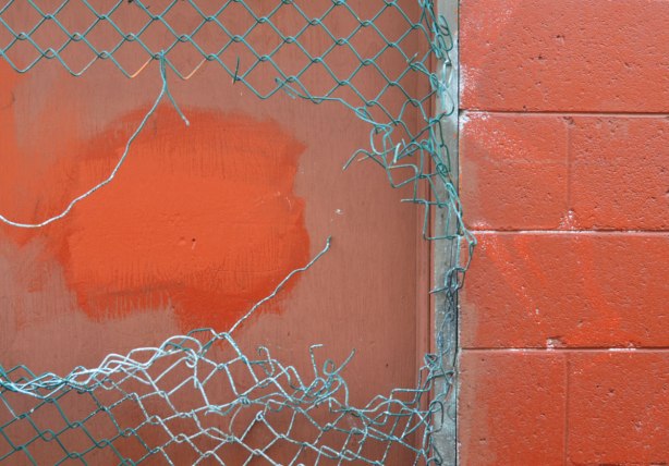A broken vinyl covered chainlink fence in front of a red wall that is part concrete block and part wood