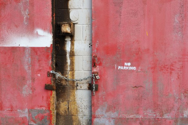 part of two old rusty and messy red doors on an old white building. There is a chain between the doors. The door on the right has the words No Parking stencilled onto it. 