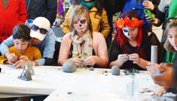Two girls with day of the dead face paint on are making clay skulls. A young boy is also at the table making a skull, his mother is helping him. 