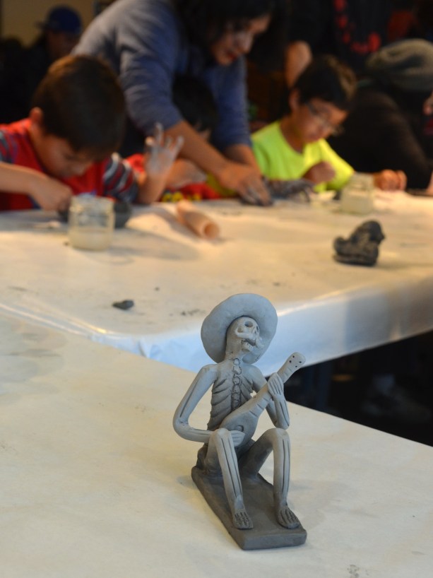 A small figurine made of a clay of a skeleton wearing a sombraro and playing a guitar is in the foreground, kids making clay skulls at a table are in the background. 