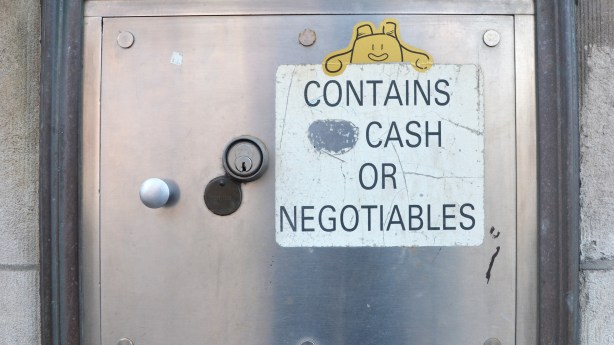 A little yellow sticker on a bank night deposit box, looks like a little smiling creature looking over the sign that says contains no cash or negotiables, except that the word no has been scratched out