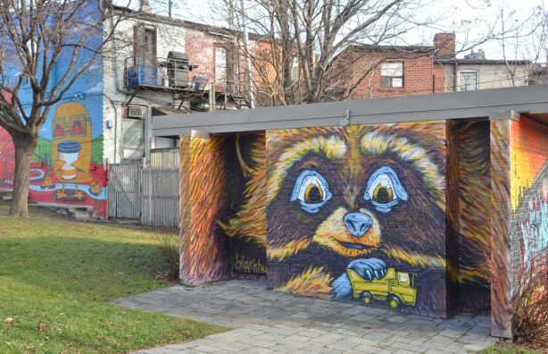 mural by blackburn of a raccoon, or similar creature, playing with a yellow toy truck, painted on the side of a building in a park