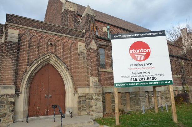 An old brick church at a corner. It is now empty. There is a large sign in front of it advertising Stanton developments. 