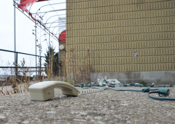 receiver of an old beige phone with its coiled wire leading to base of phone, abandoned on a sidewalk outside an old restaurant, on the ground beside a broken green wire string of Christmas lights. 