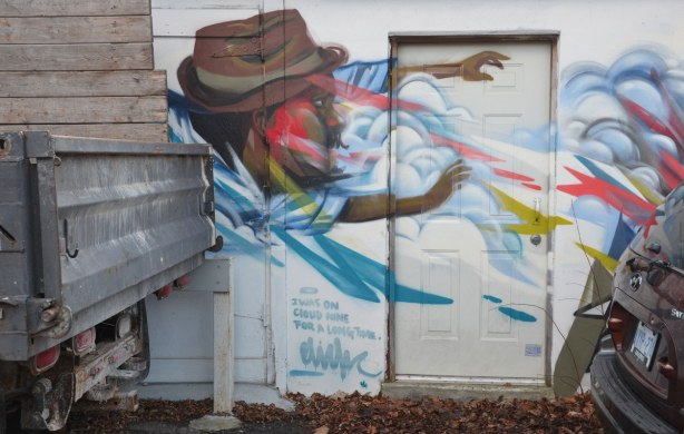 part of an elicser piece on a garage door but a car and a truck are parked in front of the rest. A man in a brown hat is blowing against a door, streams of wind coming from his mouth and clouds are around him, words say I was on cloud nine for a long time.