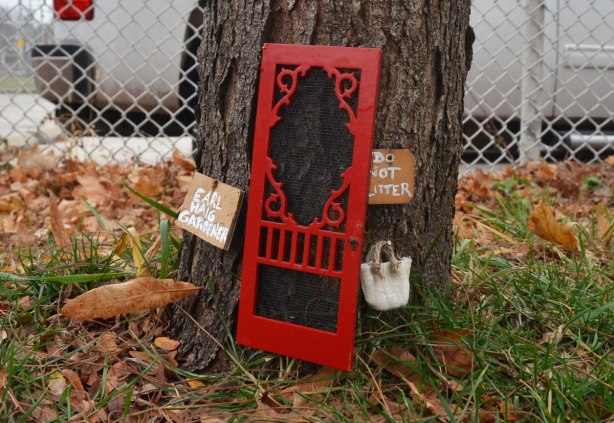 A little red screen door, fairy door, at the base of a tree with two little signs. One sign says Earl Haig gardener and the other says Do not litter.