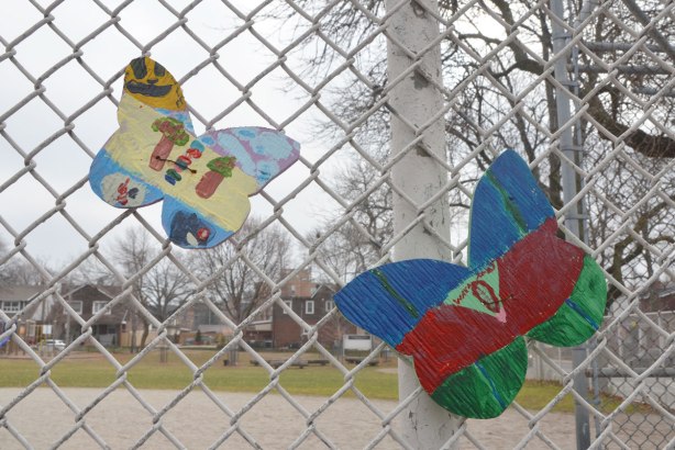 two wood butterfly shapes that have been hand painted by kids and then attached to a chain link fence around a school playground. 