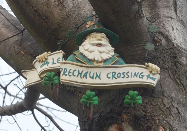 Up in a tree, a decoration with a leprechaun holding a broken sign that says leprechaun crossing. 
