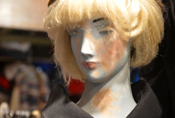 head of an old mannequin with blond wig, one eye is missing, and the covering on her is wearing off. 