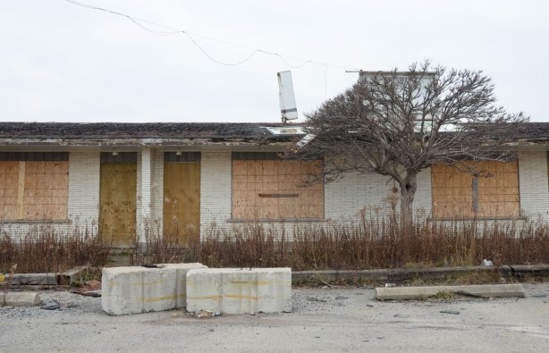 boarded up doors and windows on an old motel, the sign on the roof is broken and part has fallen to the ground. 