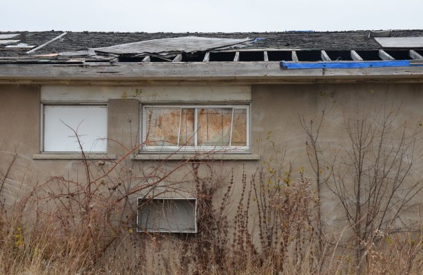 back of motel, some roof shingles missing, inside of windows has been covered, weeds overgrown, 