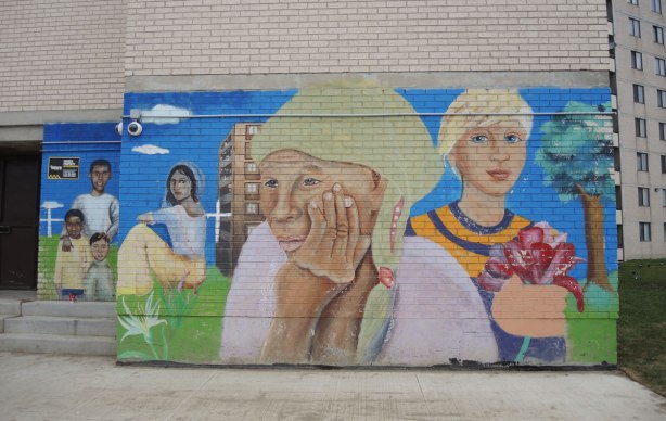 part of a mural on the lower floor of a grey brick apartment building, women, one holding flowers, one resting her head on her hand, one sitting on the grass.