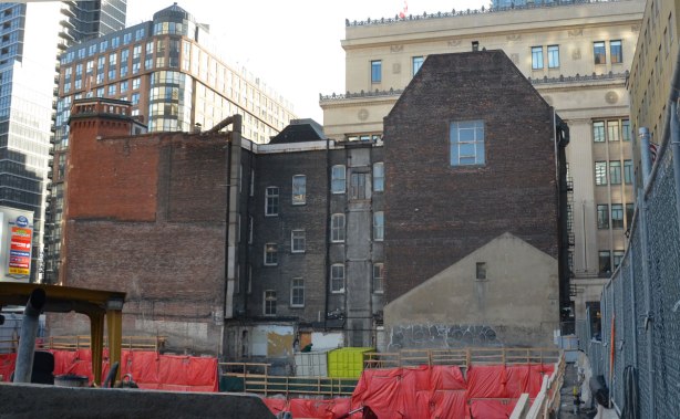 back of a large four storey brick building behind an open hole construction site, taller buildings in the background (College Park)