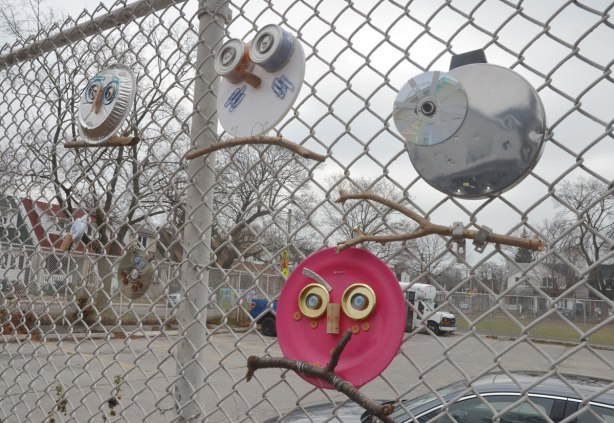 four owls made of recycled goods, foil pie plates, CDs, bottle tops, there feet are wrapped around twigs and they are attached to a chainlink fence 