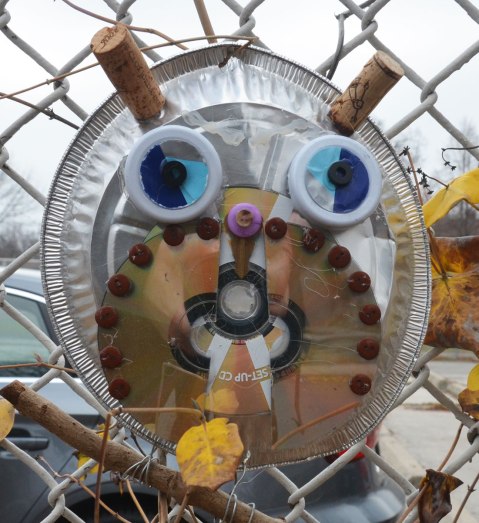owls made of recycled goods, foil pie plates, CDs, bottle tops, there feet are wrapped around twigs and they are attached to a chainlink fence - two corks for horns