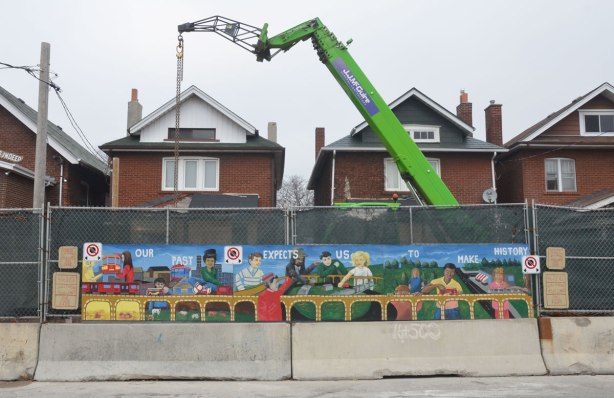 mural in front of a construction site, the tops of two brick houses are visible behind the fence, a large green crane is working at the site