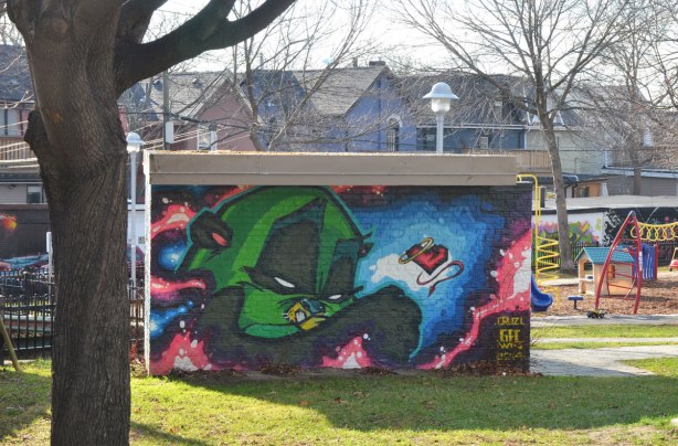 side of a small building covered with a mural of a green bird-like character, playground and houses in the background