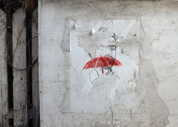 A drawing of a man on white paper pasted to a concrete wall. A red stenciled umbrella is on top of his face. A white picket fence is beside the wall. 