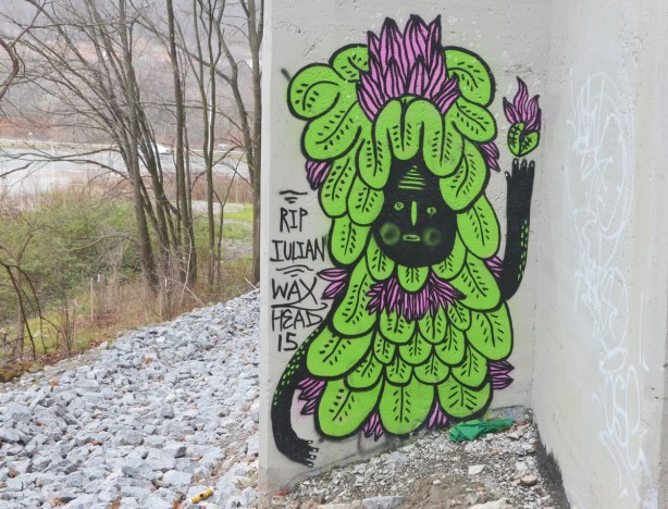 graffiti on concrete bridge supports - creature with black face and covered in green leaves, with a few purple petals on top of the head. words, RIP Julian Waxhead