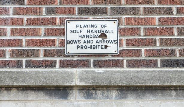 old sign on the exterior wall of a school that says: Playing of golf, hardball, handball, bows and arrows prohibited