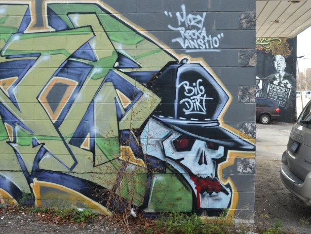 street art in an alley, a white skull with a black baseball cap. Cap says Big Dirt on it. Words above the skull are Mary Recka Pantilo (could be Mozy not Mary)