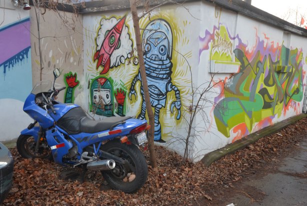A blue motorcycle is parked in front of a street art painting of three spacemen creatures