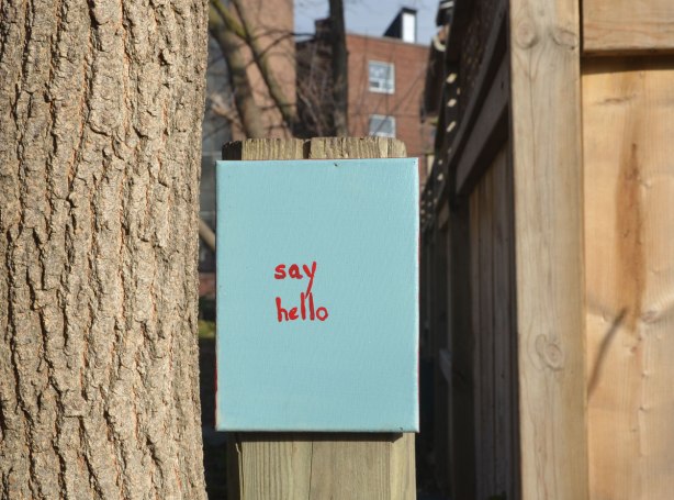 A small canvas painted turquoise and the words say hello written in small red letters, attached to a fence in a lane