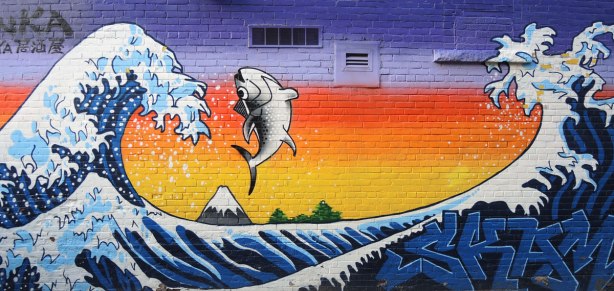 mural based on a Japanese picture called the Great Wave. In it there is a fish jumping with the wave. Mural on the side of a restaurant in a lane.