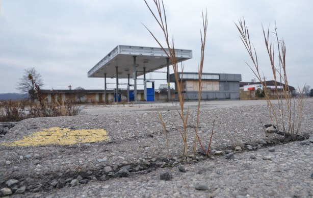 view from down low, weeds growing up through cracks in the pavement with an abandoned gas pumps and boarded up motel in the background