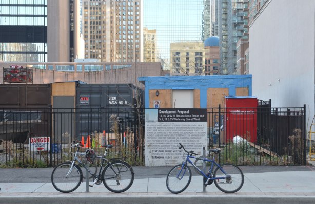 two bicycles parked on a sidealk in front of a fence that has a development proposal sign on it. Building site behind that, thena wall of skyscrapers in the background. 