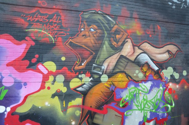 mural on an exterior wall, a monkey in old fashioned aviator's helmet and pink scarf is shouting "What's all the noise about"