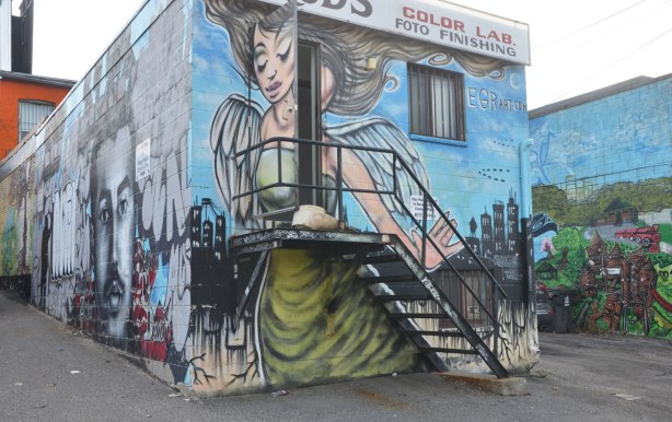 a large mural of a woman with wings that takes up the entire back of a two storey building in a laneway. There is also a metal staircase with a small porch. The door at the top is open so part of the woman in the mural opens up too. The mural is by EGR art.