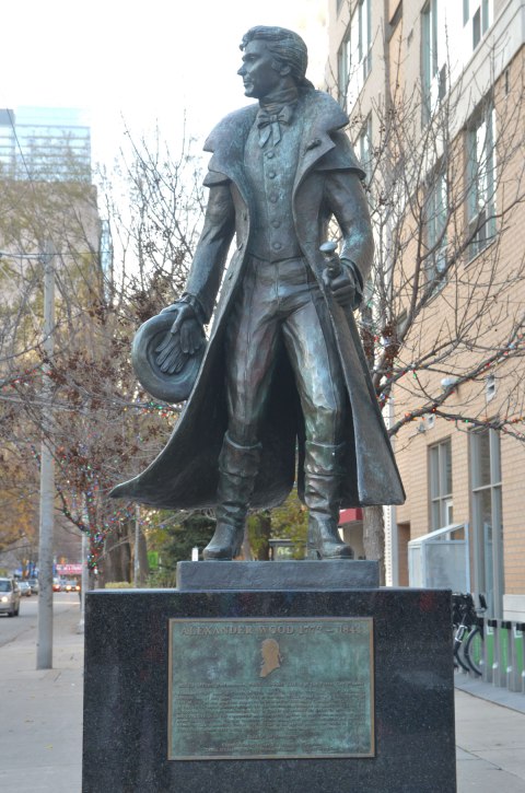 statue of Alexander Wood, a young man with long coat, and a hat in his hand. The statue is on a large square pedestal so his feet are close to eye level.  Below the statue is a plaque detailing his life as early settler of York, of being a gay, and of being involved in a scandal in 1810. 
