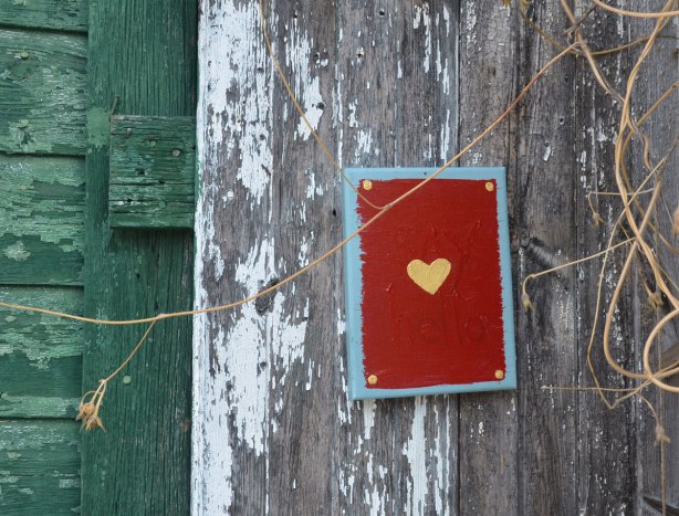A small canvas painted red and a small yellow heart painted in the middle. The words say hello are painted on it too but they are harder to see. It is nailed to an old wood fence