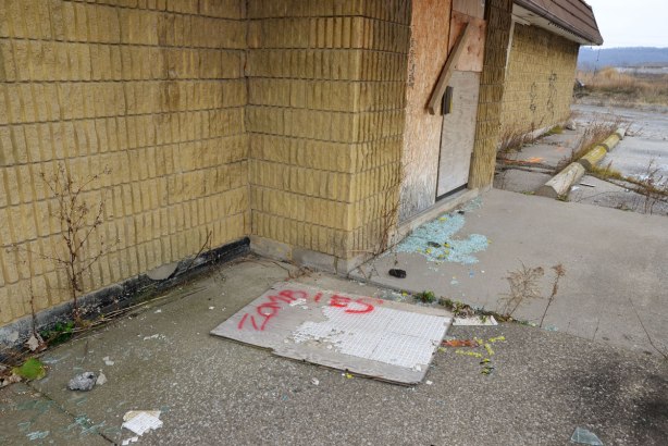 Outside the boarded up doors of a motel, broken glass, a piece of wallboard with some one inch tiles still attached on which someone has spray painted the word zombie in red