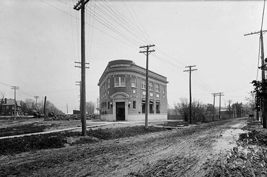 Historical black and white photo of a two sorey brick building at an intersection of two dirt roads. 