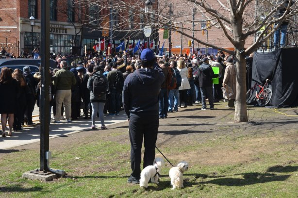 A man with two little white dogs on a leash stands in front of St. James cathedral along with a crowd of people there for Rob Ford's funeral 