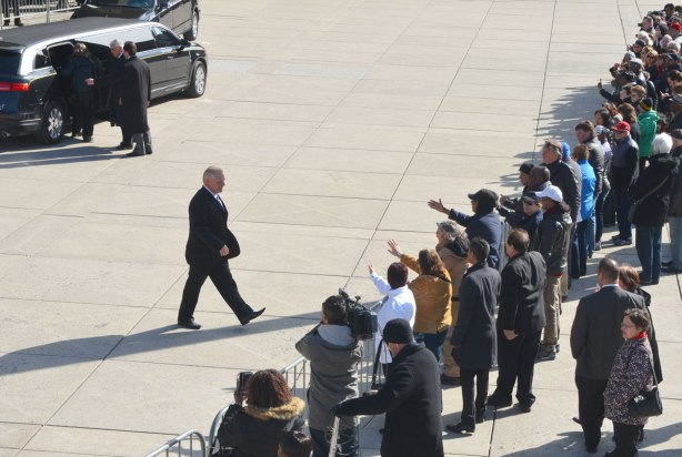 Doug Ford walks from a black limo to a crowd of people standing behind barricades in front of City Hall. They have their arms outstretched towards Ford, ready for a handshake and greeting. 