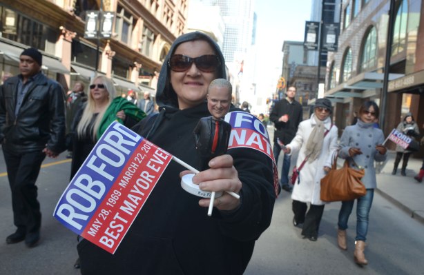 A woman in hoodie and sunglasses holds two small Ford Nation flags as well as a bobble head doll of Rob Ford as she walks in his funeral procession down Yonge Street