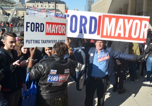 A middle aged man holds a banner that reads Ford Mayor over his head, beside him is a woman also holding a Ford Mayor sign. On the back of her jacket are a number of stickers in support of Ford 