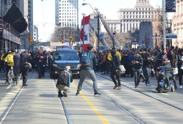 A funeral procession for Rob Ford passes along Queen Street on its way to St. James cathedral, photographers are in front, a police guard is walking beside it.