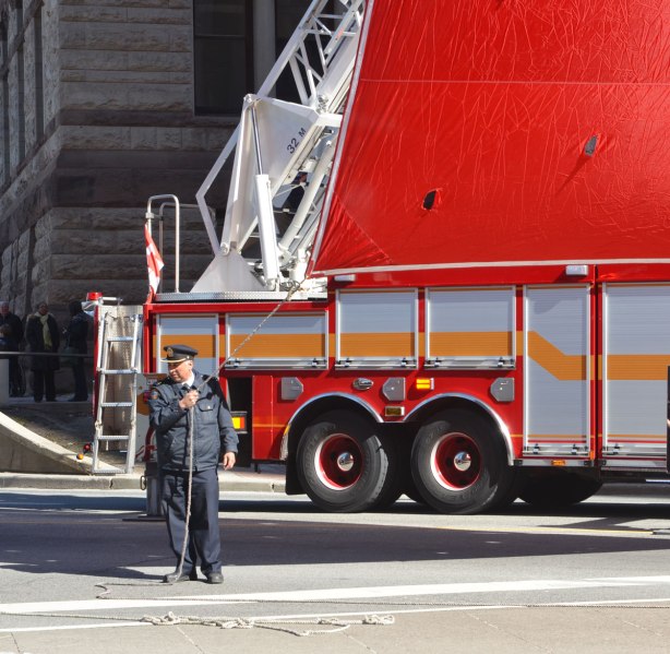A fireman holds a rope that is attached to the corner of a very large Canadian flag. A firetruck is behind him
