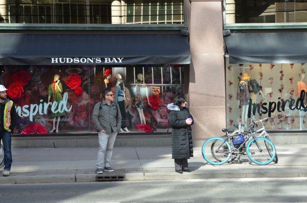 Three people stand on the sidewalk in front Hudsons Bay store windows. A man with a hard hat, a man with hands in his pockets, and a woman in long black coat. A couple of bikes are parked there too. The theme of the store windows is Inspired. 