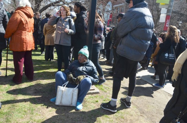a young person sits on the grass, resting against a tree while other people stand around